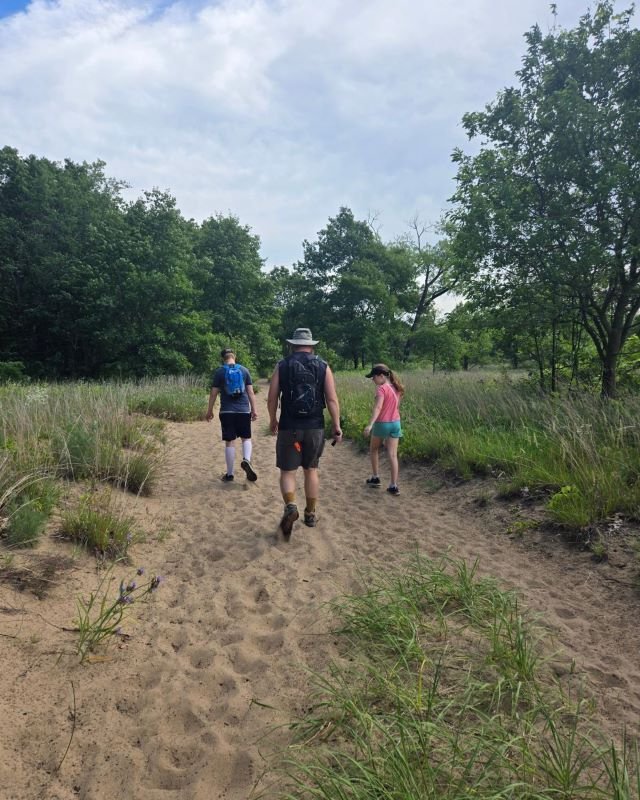 A father, son, and daughter hiking in sand next to a forest at Indiana Dunes National Park. Their back are to the camera.
